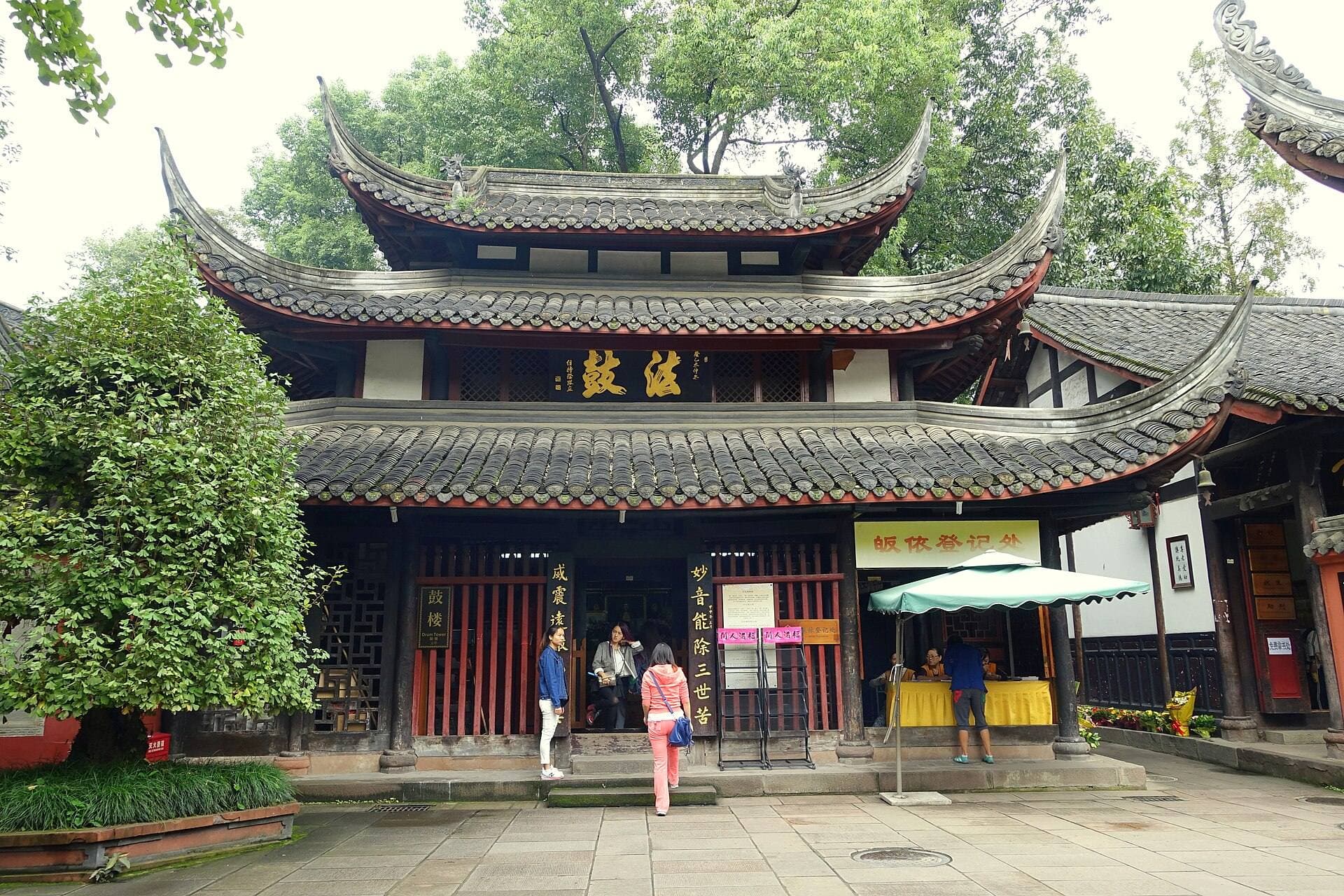 A two-tiered hall with dark upswept roofs and red wooden doors at Wenshu Monastery in Chengdu, surrounded by green trees