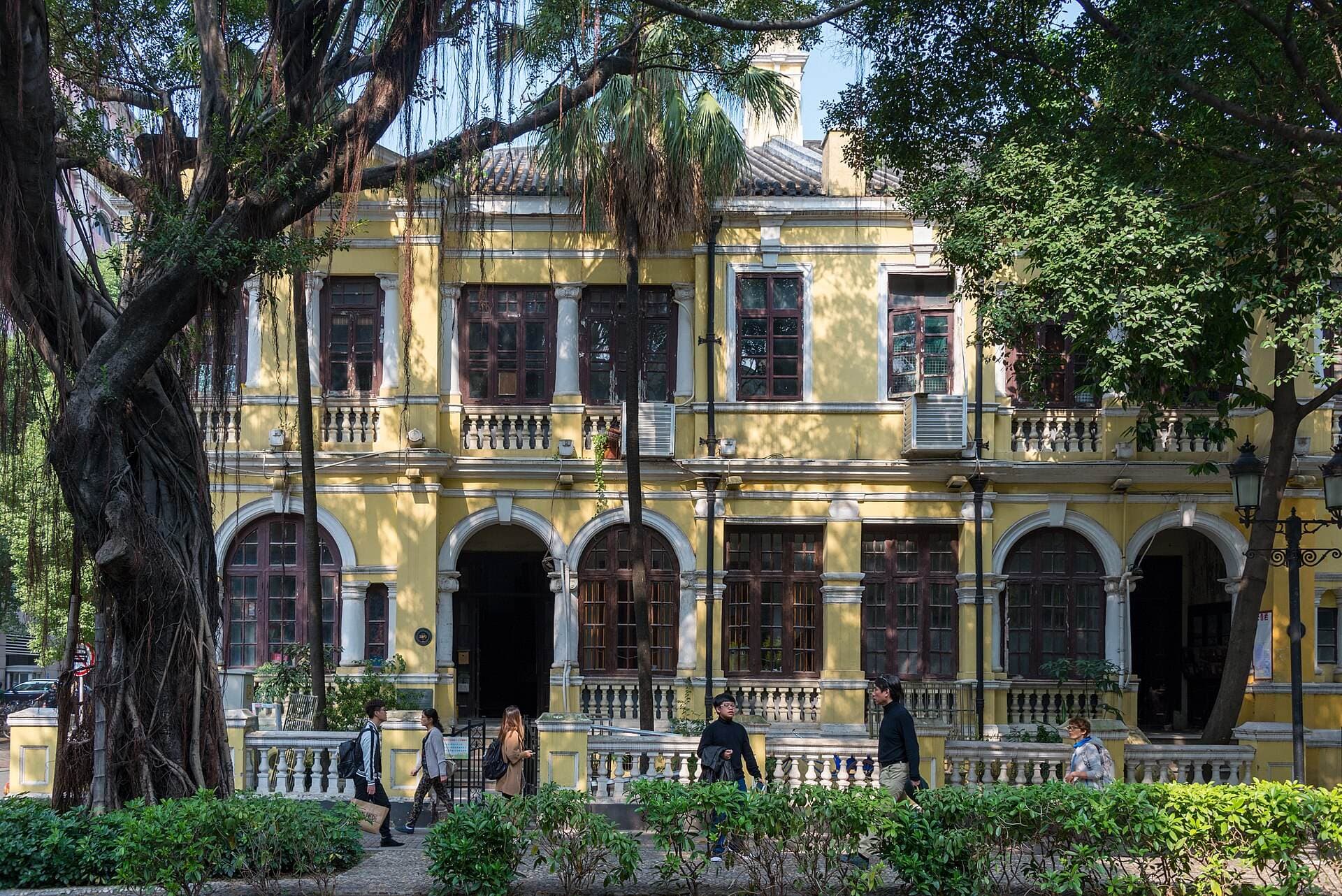 A yellow colonial-era building with arched windows and balconies on Shamian Island in Guangzhou, shaded by large banyan trees