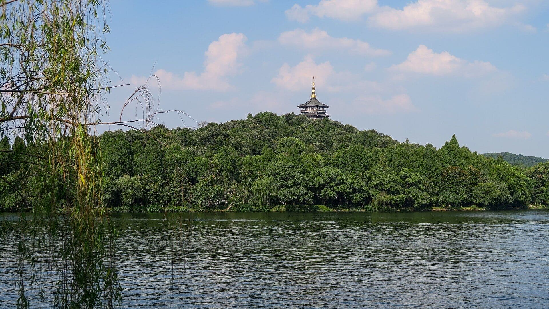 Leifeng Pagoda on a wooded hill across the calm water of Hangzhou's West Lake, framed by a weeping willow