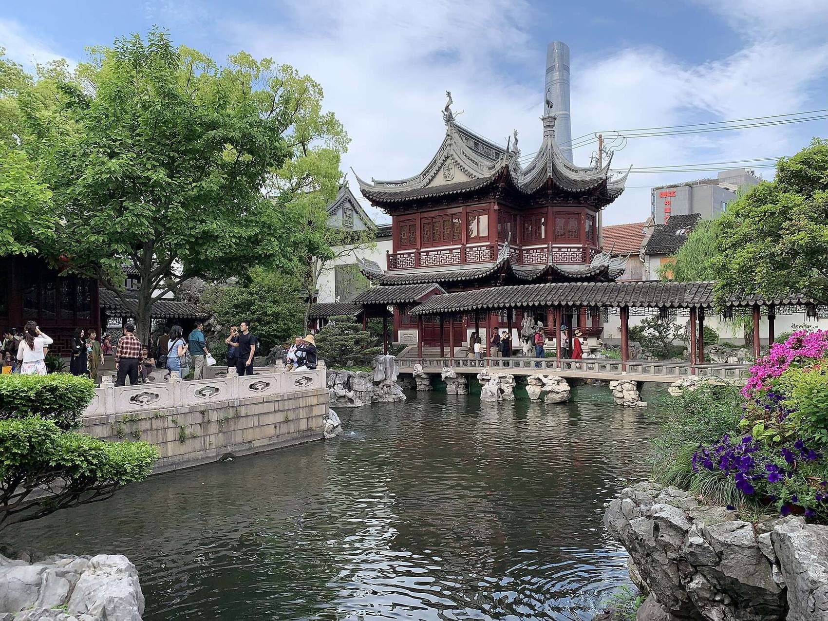 A classical pavilion overlooks a pond in Shanghai's Yu Garden, framed by trees and purple flowers, with the Shanghai Tower in the distance