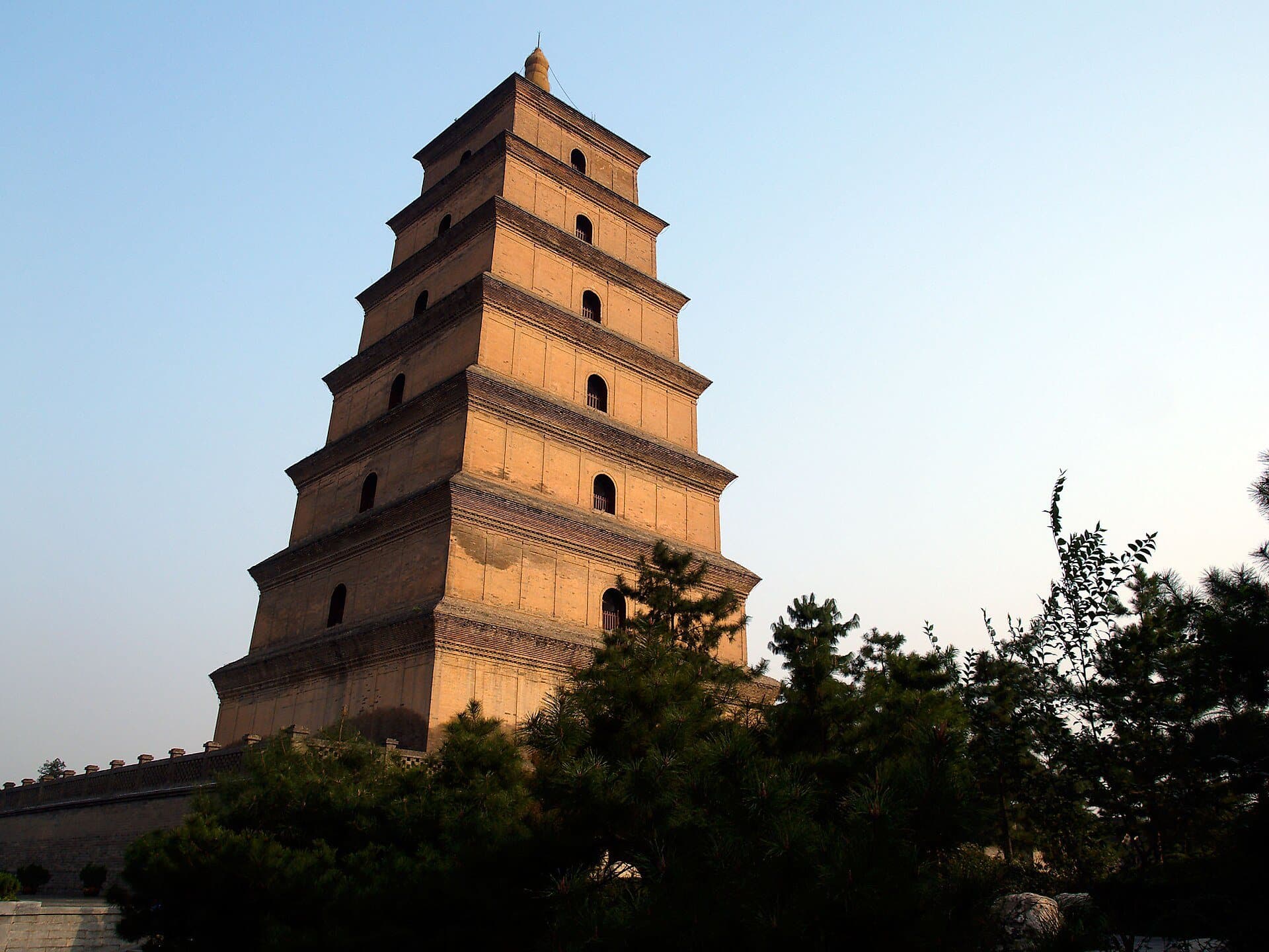 The tiered brick Giant Wild Goose Pagoda in Xi'an glows in warm sunlight above dark pine trees against a clear sky