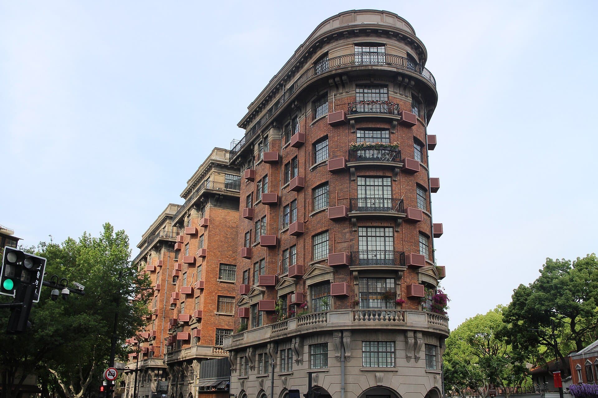 Shanghai's wedge-shaped, red-brick Wukang Mansion with wrought-iron balconies, among green plane trees under a pale blue sky