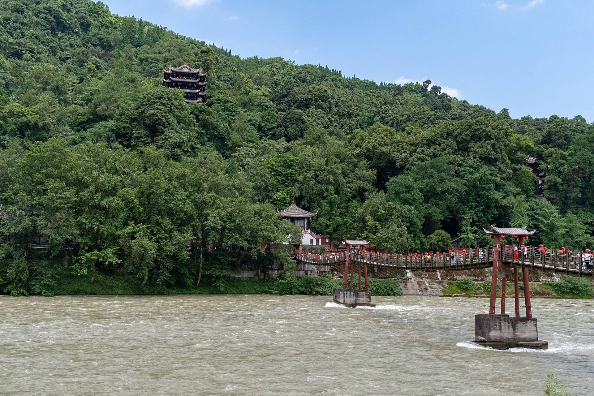 The red-pillared Anlan Suspension Bridge spanning the fast Min River at Dujiangyan, below a forested hill with a pavilion