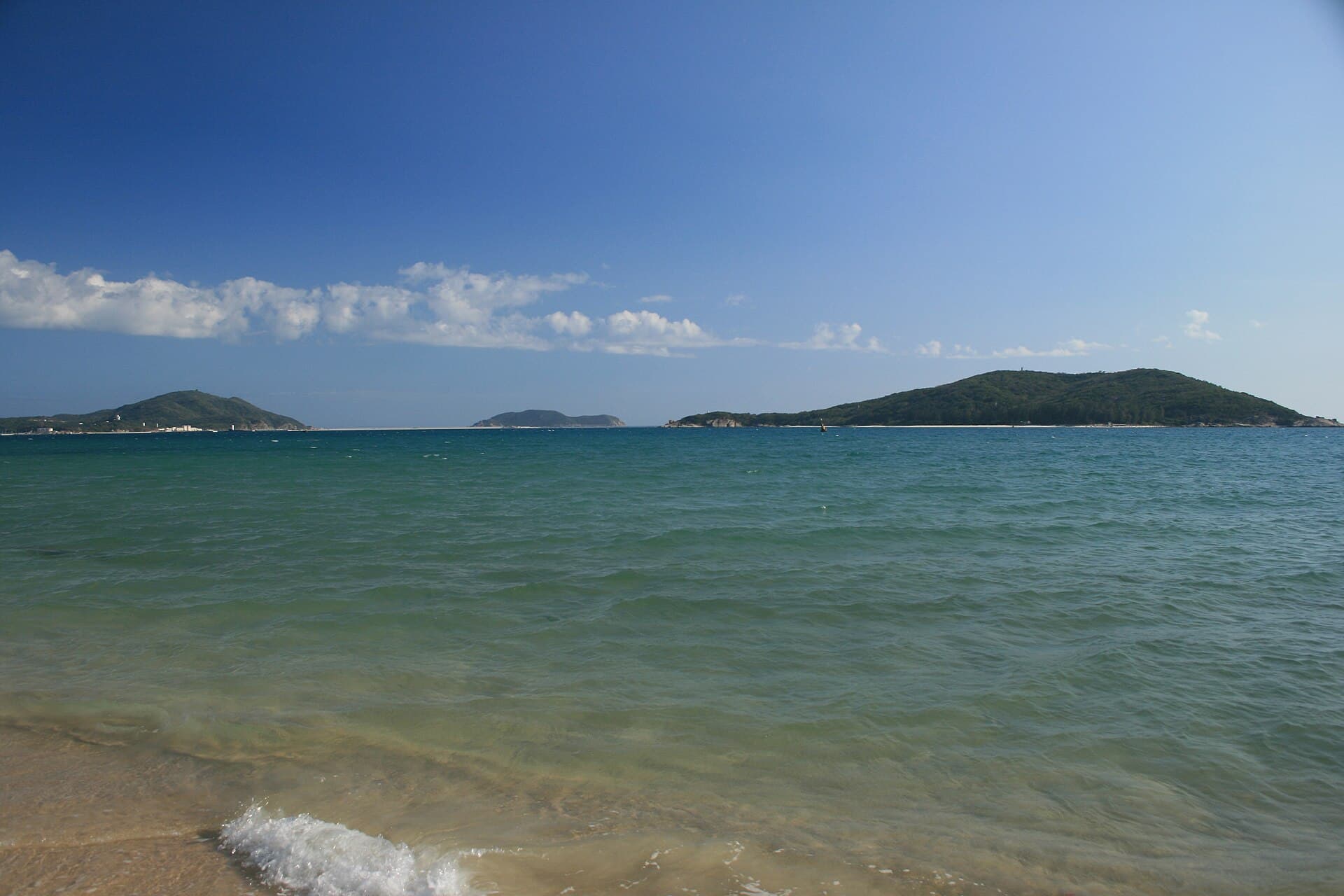 Clear turquoise water lapping a sandy shore at Yalong Bay, Sanya, with green islands on the horizon