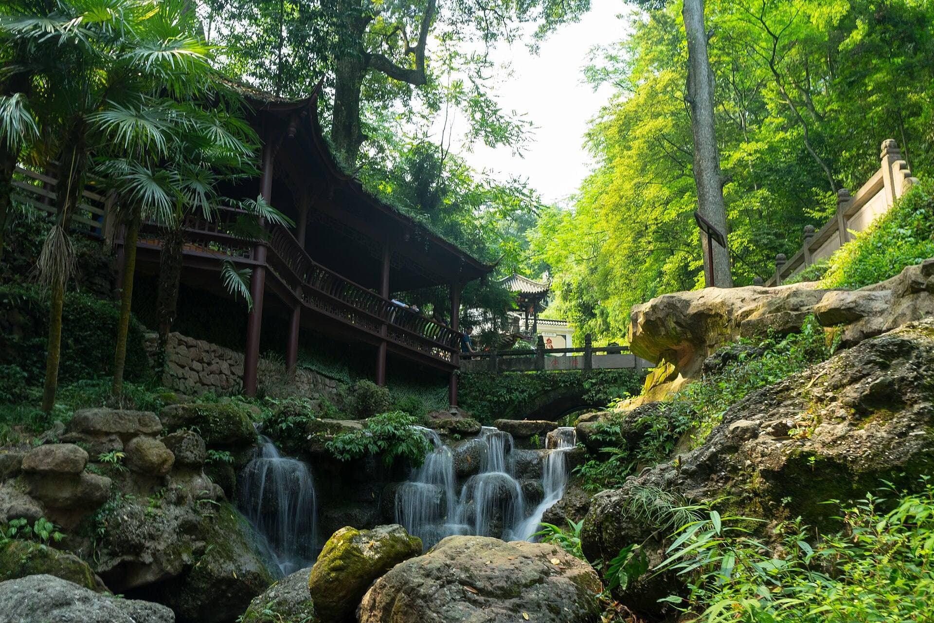 A small waterfall tumbles over mossy rocks beneath a wooden gallery and stone bridge in the forest of Mount Qingcheng