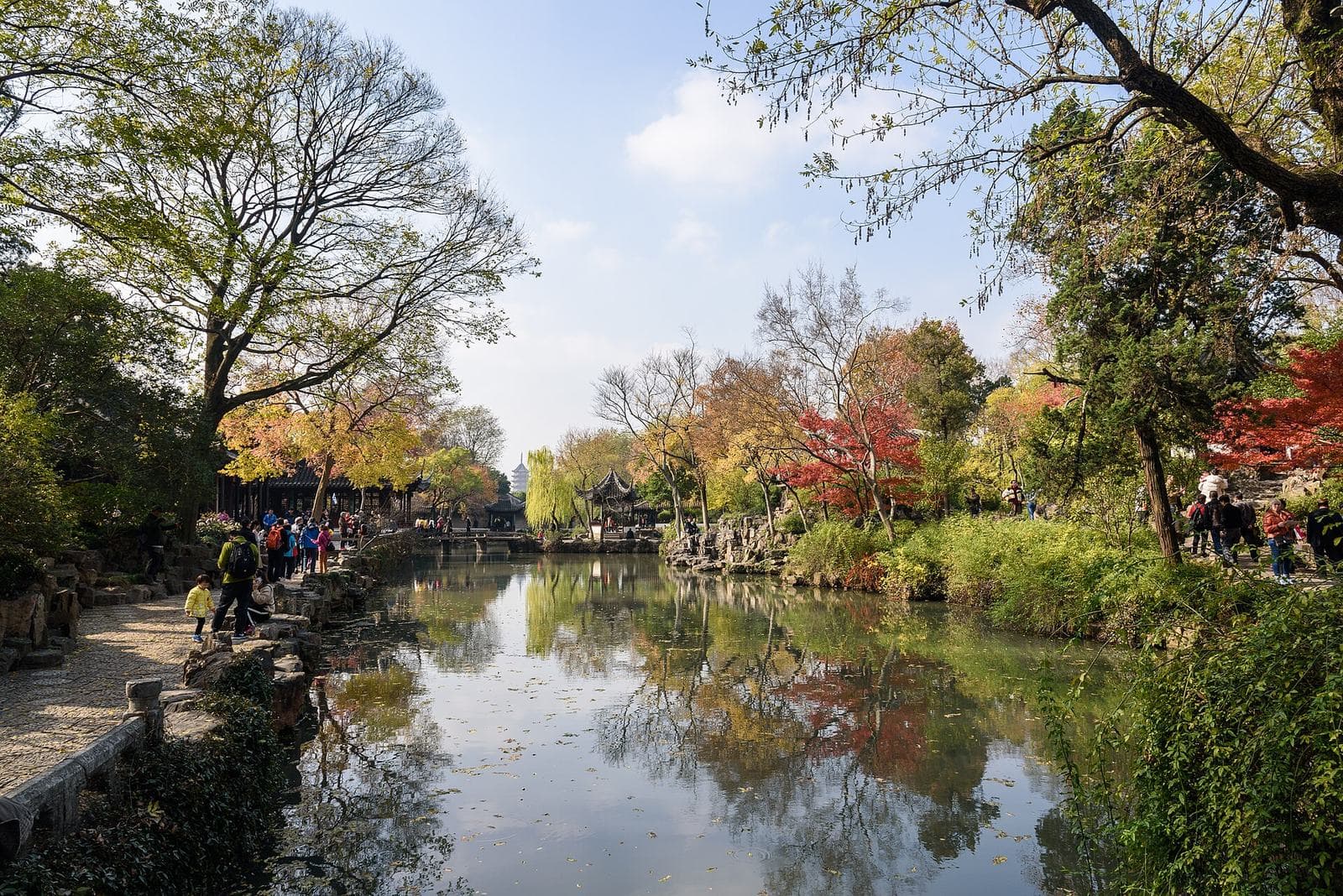 A still pond in the Humble Administrator's Garden in Suzhou, reflecting autumn trees, red maples, a pavilion and a distant pagoda
